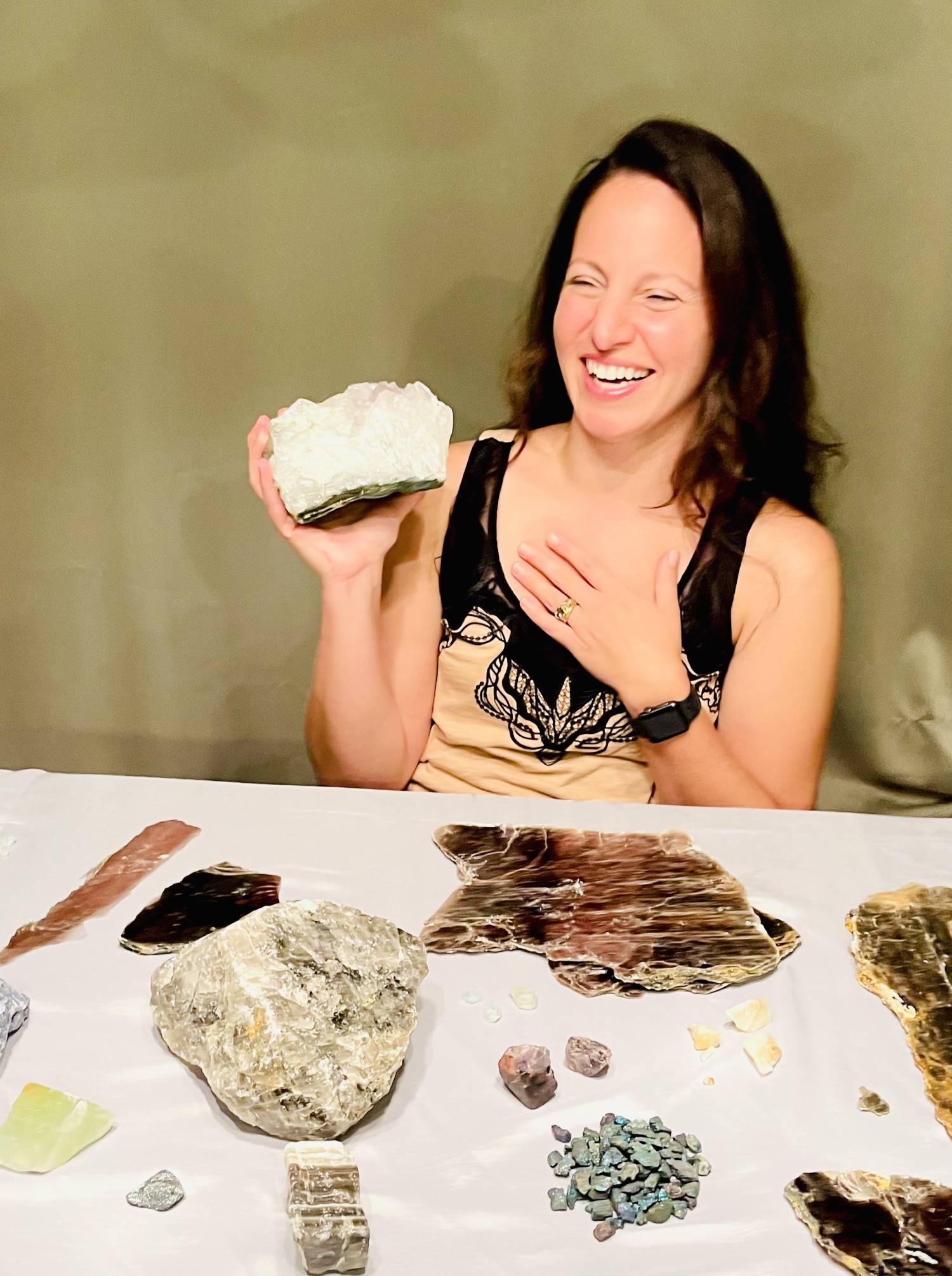 Tammy Campbell sitting at a table with gemstones and minerals, holding a large uncut gem and laughing.