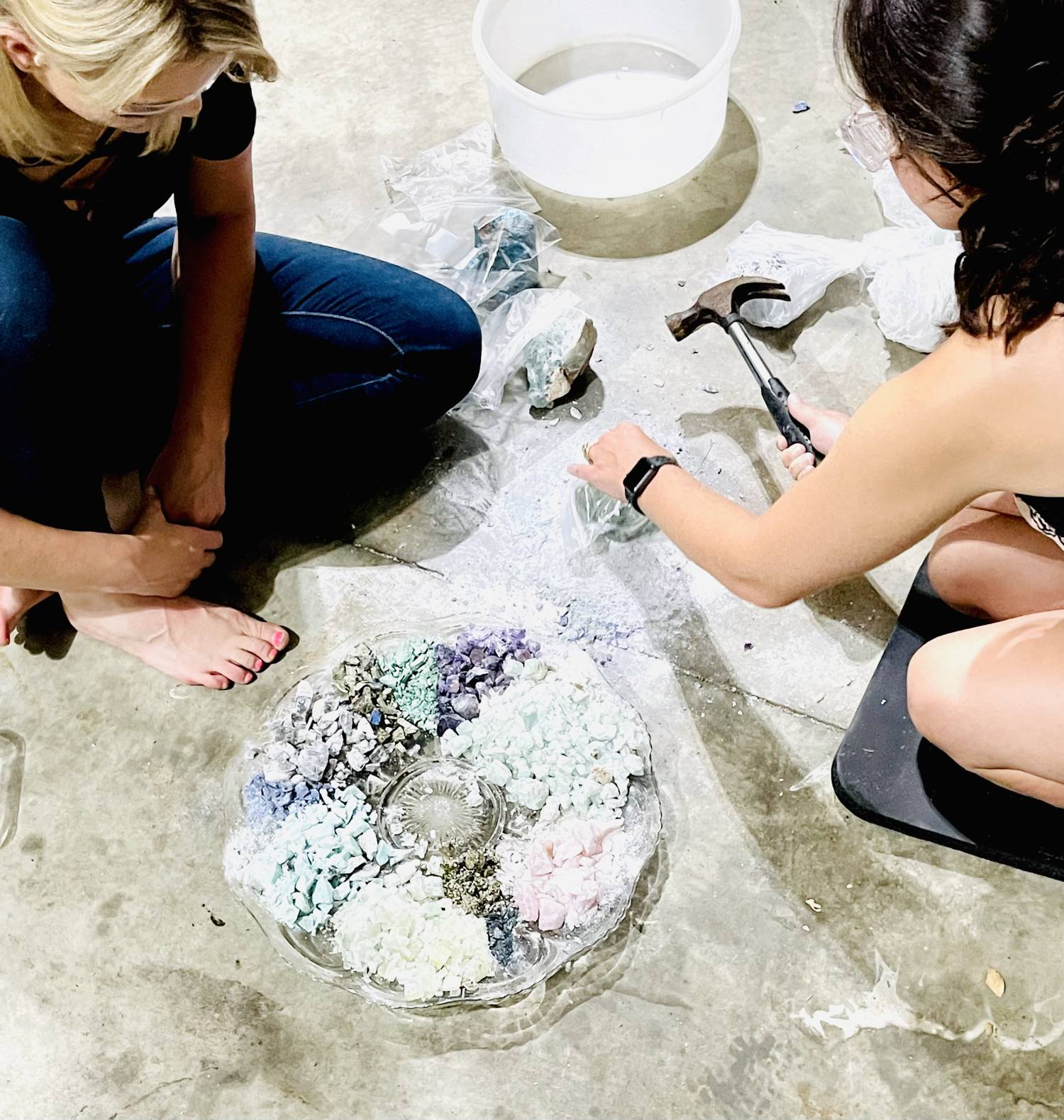 Photo of Tammy Campbell smashing gemstones with another person who is blonde and barefoot. They are both sitting on the floor next to a platter of smashed gemstones.