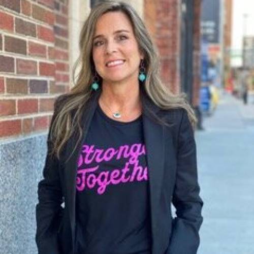 Photo of Flora Migyanka wearing a shirt with pink lettering "Strong Together" standing on a street next to a brick wall.